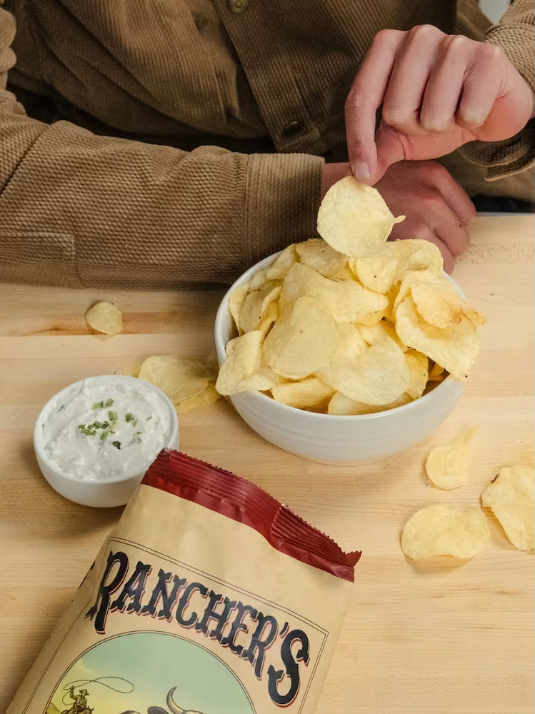 Person dipping a chip into a bowl of rancher's chips with a bag of rancher's chips and a small bowl of dip on a wooden surface.